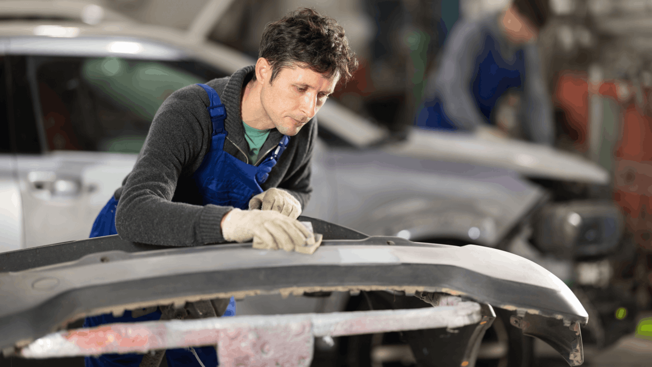 Auto body parts technician in blue overalls sanding and preparing vehicle panel for repair in professional collision shop