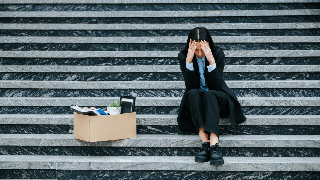 Worker sitting on steps after job loss holding head in hands next to box of belongings