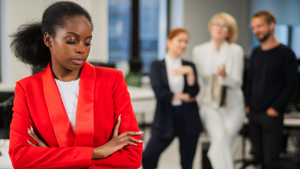 Black woman standing alone with arms crossed while coworkers whisper behind her depicting racial discrimination