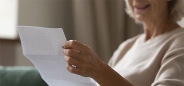 Close up wrinkled female hands holding paper document. Smiling happy middle aged senior woman reading letter, feeling excited by good news